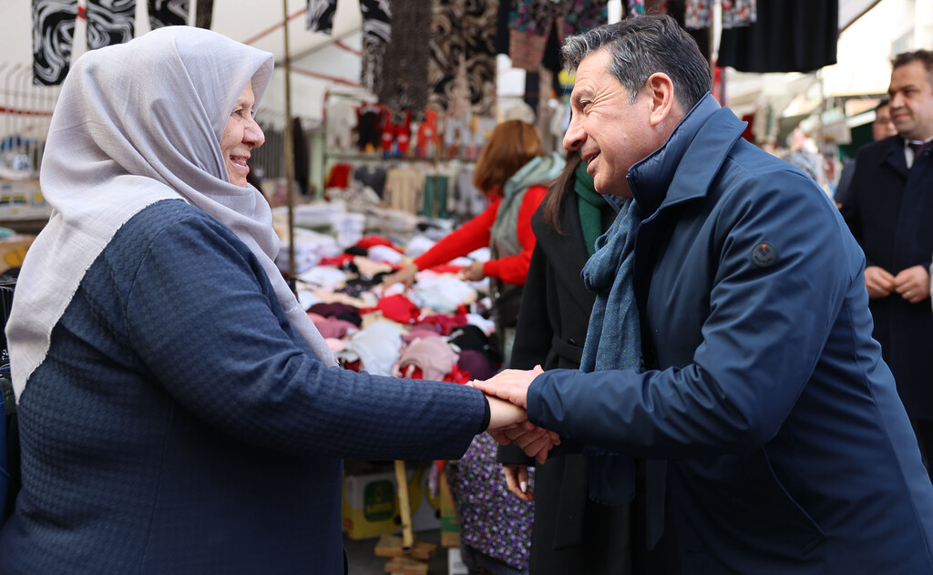 Muğla Büyükşehir Belediye Başkanı Ahmet Aras, bir semt pazarında gülümseyen başörtülü bir kadınla tokalaşıyor ve her iki eliyle kadının elini tutarak sıcak bir şekilde sohbet ediyor.