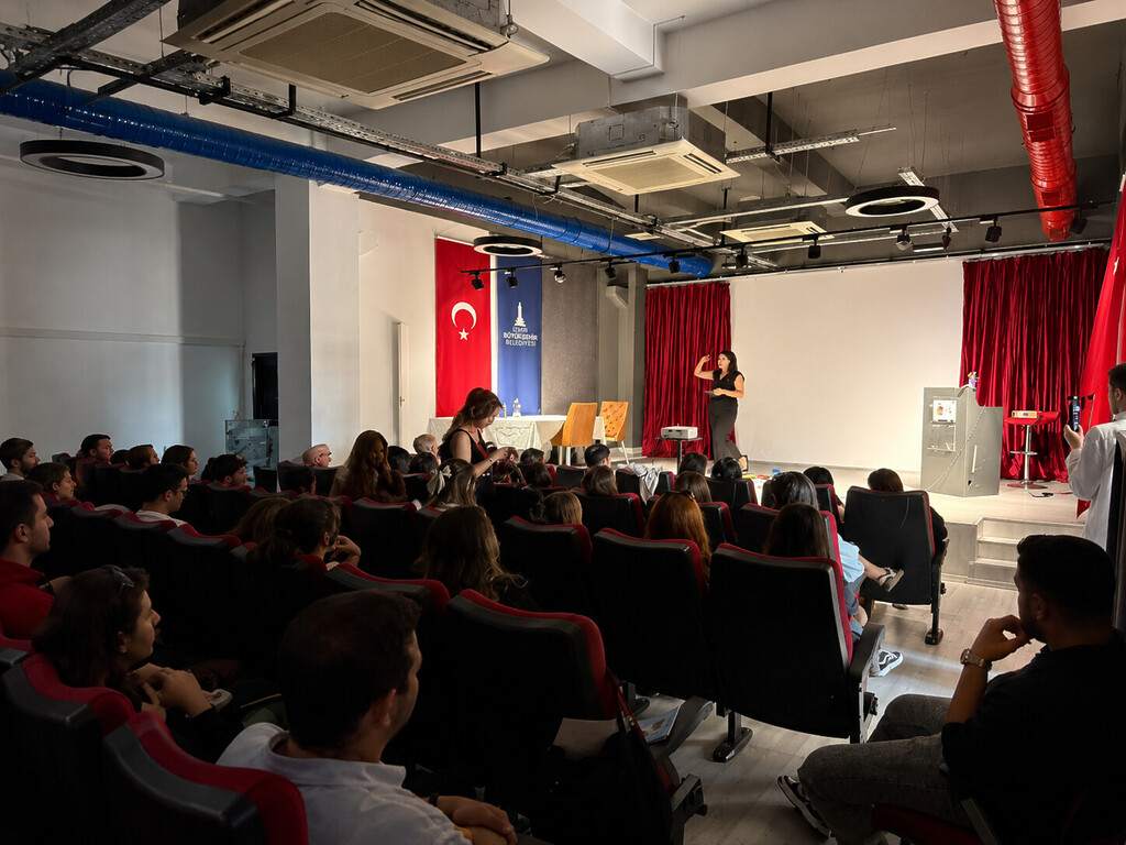İzmir Büyükşehir Belediyesi tarafından düzenlenen ADİS seminerinde, bir uzman sahnede dinleyicilere sunum yapıyor. Salonda dinleyiciler koltuklarda oturuyor; arka planda Türk bayrağı ve belediye flaması yer alıyor.